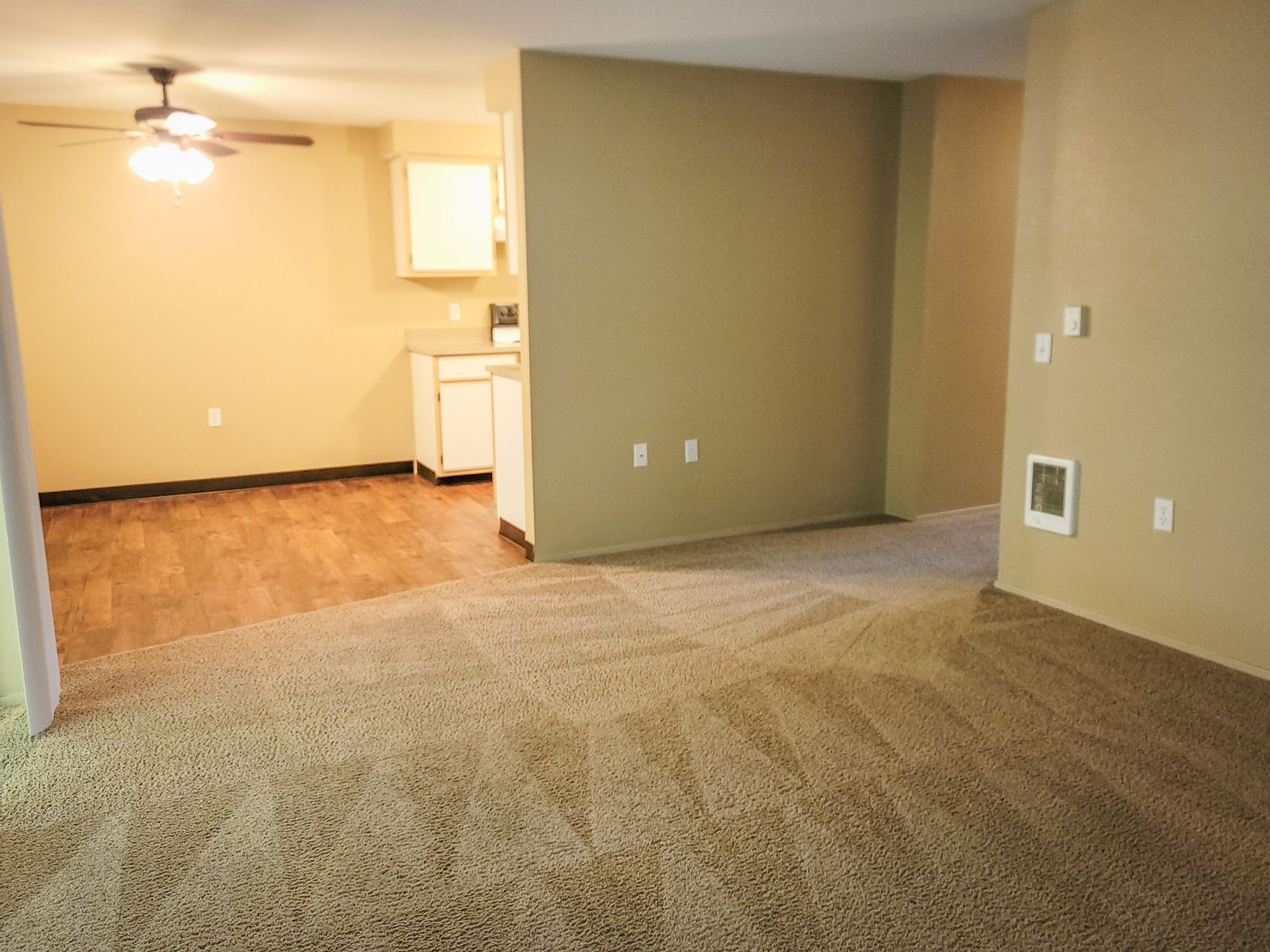 Empty carpeted living room with a small kitchen area and beige walls, viewed from a doorway