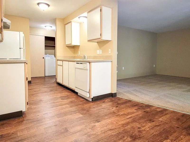 Empty apartment interior with kitchen, wood floors, and carpeted living area