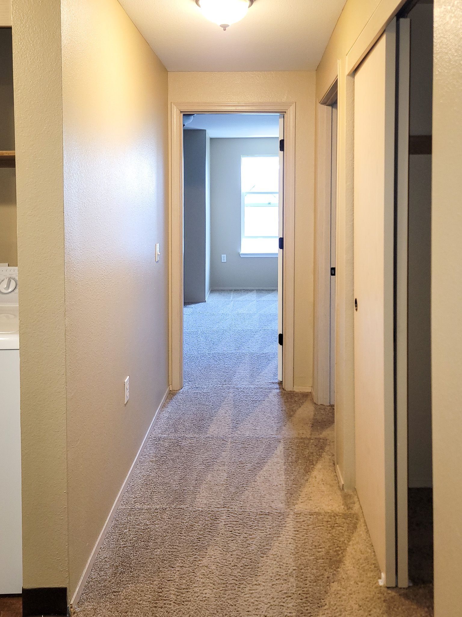 Narrow beige hallway with carpet leading to a bright room at the end.