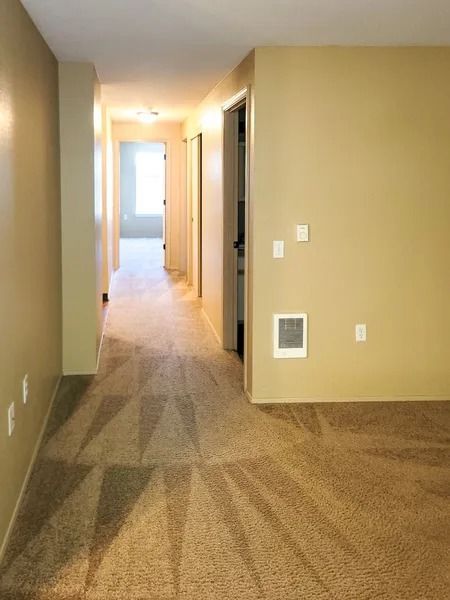 Empty beige hallway with carpet, vents, and an open doorway leading to a bright room.