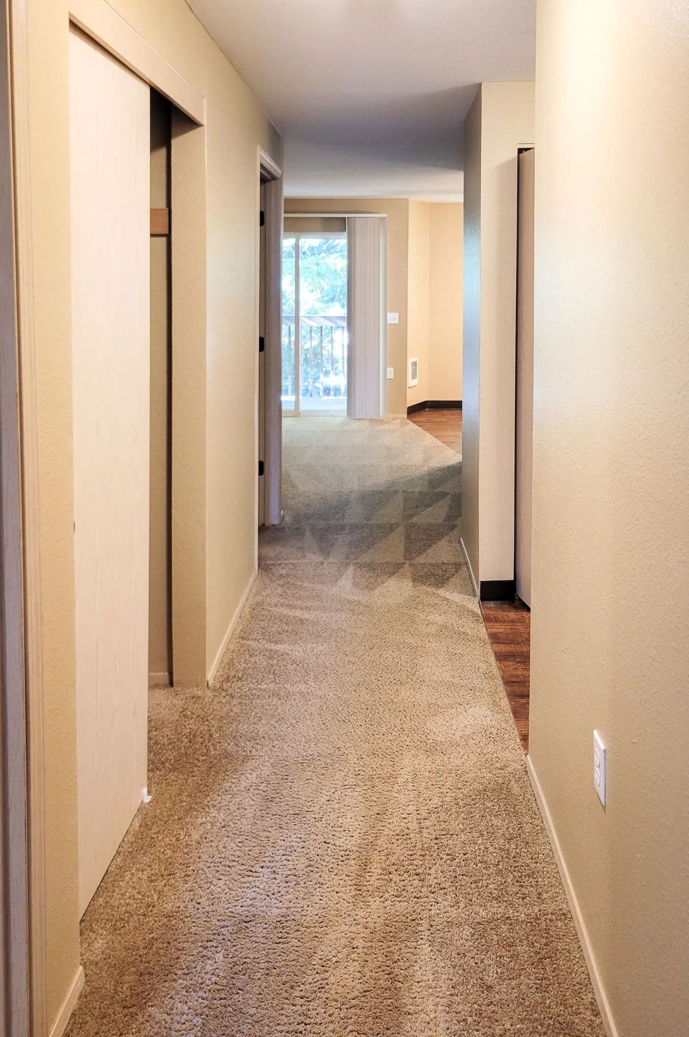 Narrow carpeted hallway in a home with beige walls and doors, leading to a bright window at the end.