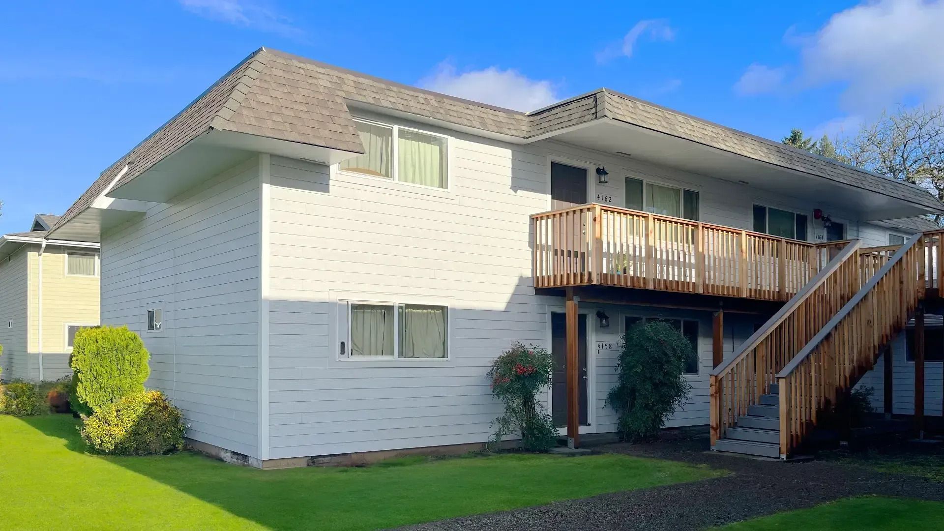 White two-story apartment building with wooden balconies, exterior stairs, and a green lawn.