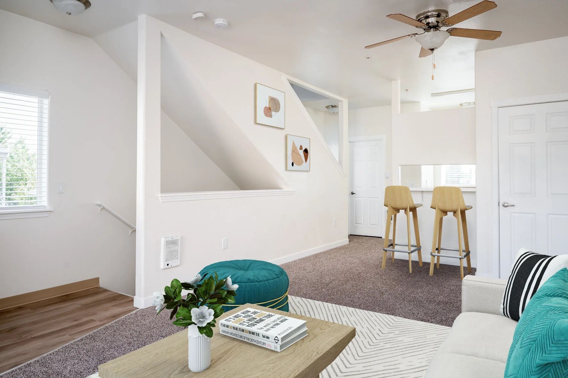 Bright living room with white walls, teal sofa, coffee table, staircase, and breakfast bar stools near a kitchen.