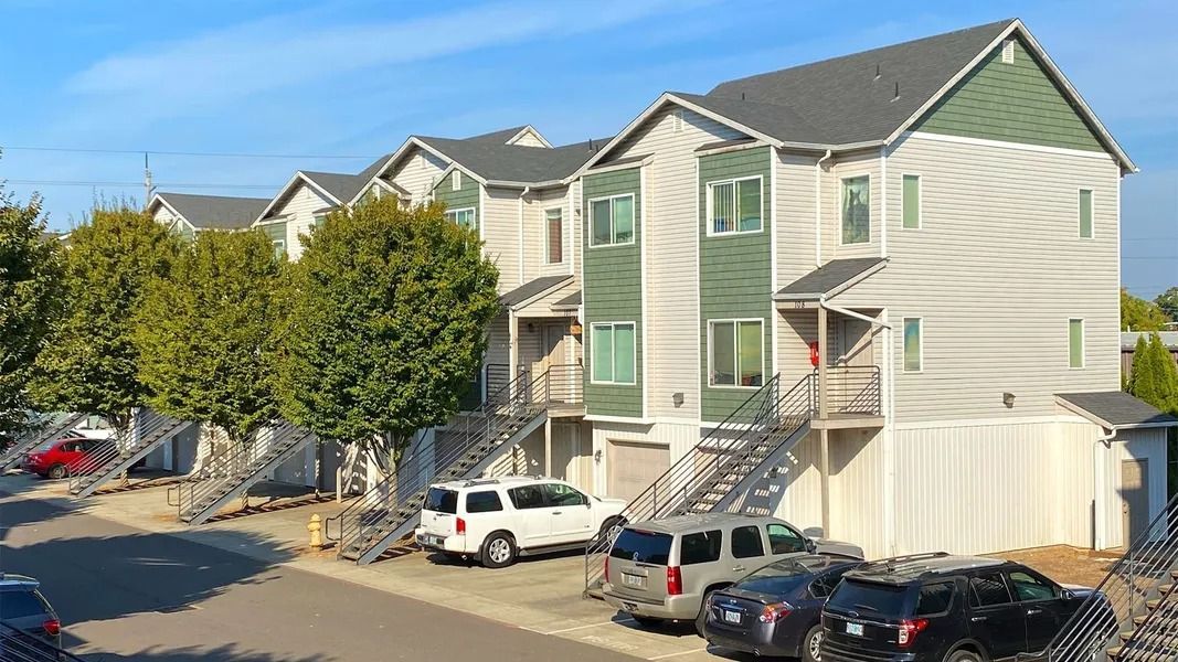 Apartment complex with green siding, external stairs, parked cars, and trees along a sunny street.