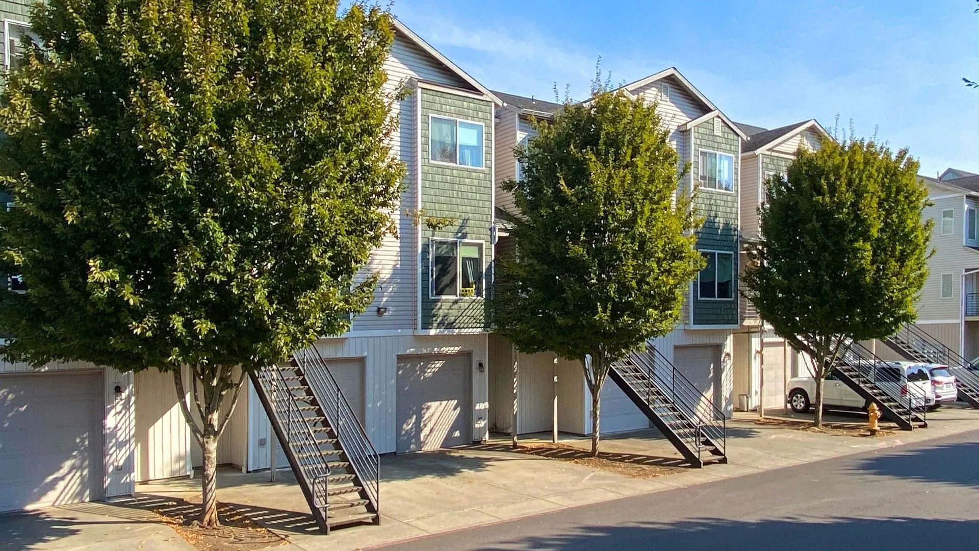 Row of townhouse exteriors with white garages, black staircases, and trees along a sunny street