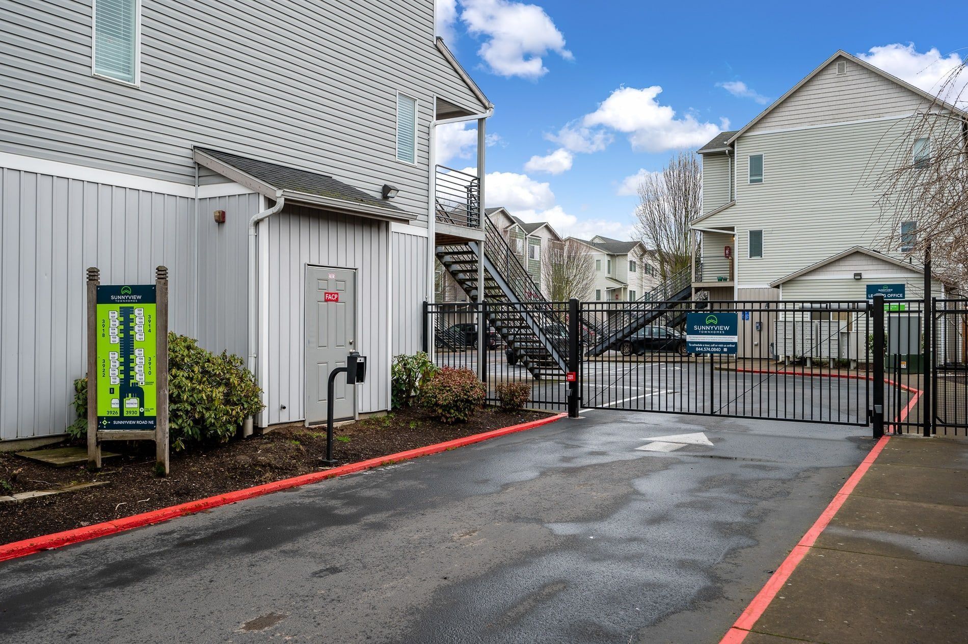 Gated apartment parking lot with wet pavement, black metal fence, and white buildings under a cloudy sky