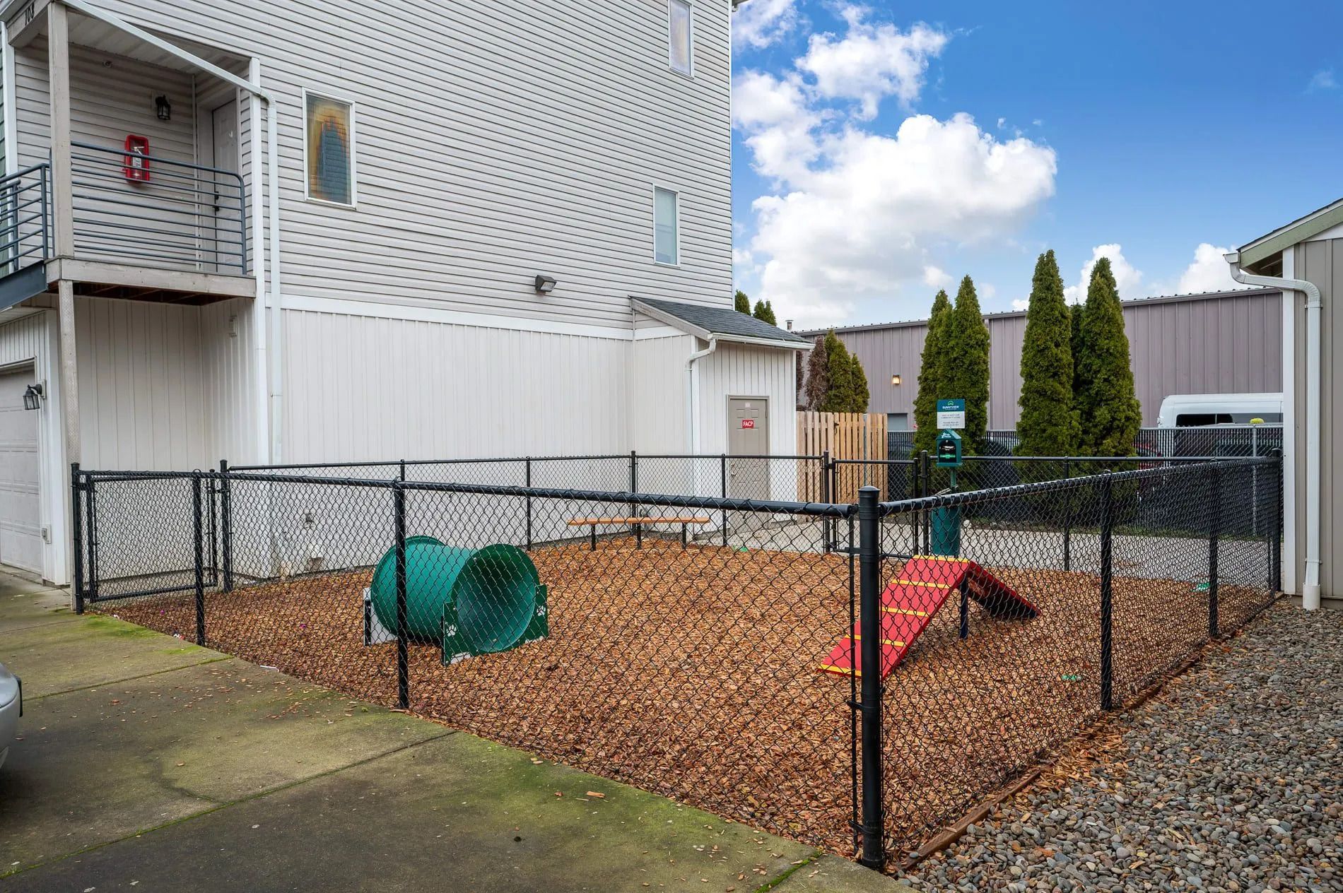 Small fenced backyard dog run with wood chips, green tunnel, and red ramp beside a white building