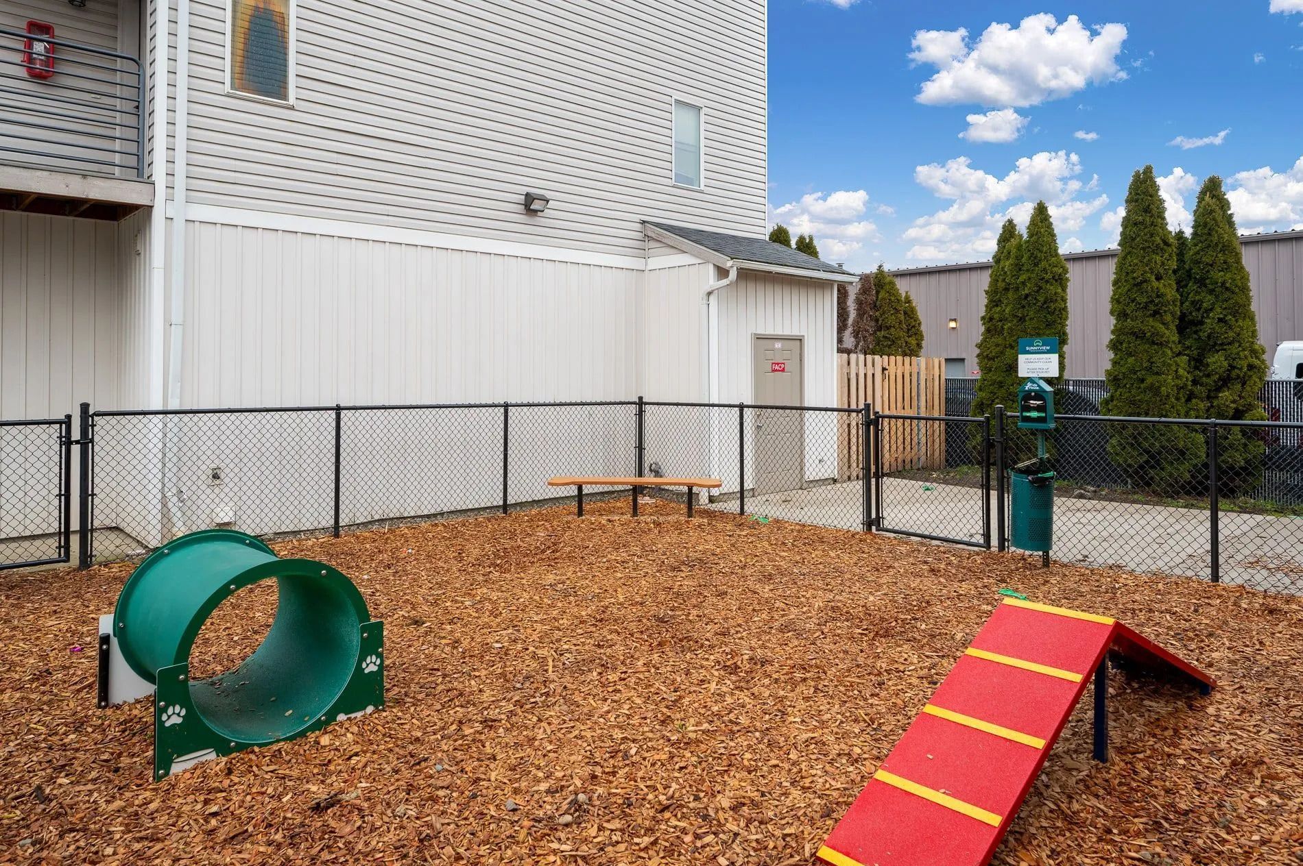 Backyard playground with wood chips, green tunnel, red ramp, and fenced play area beside a house