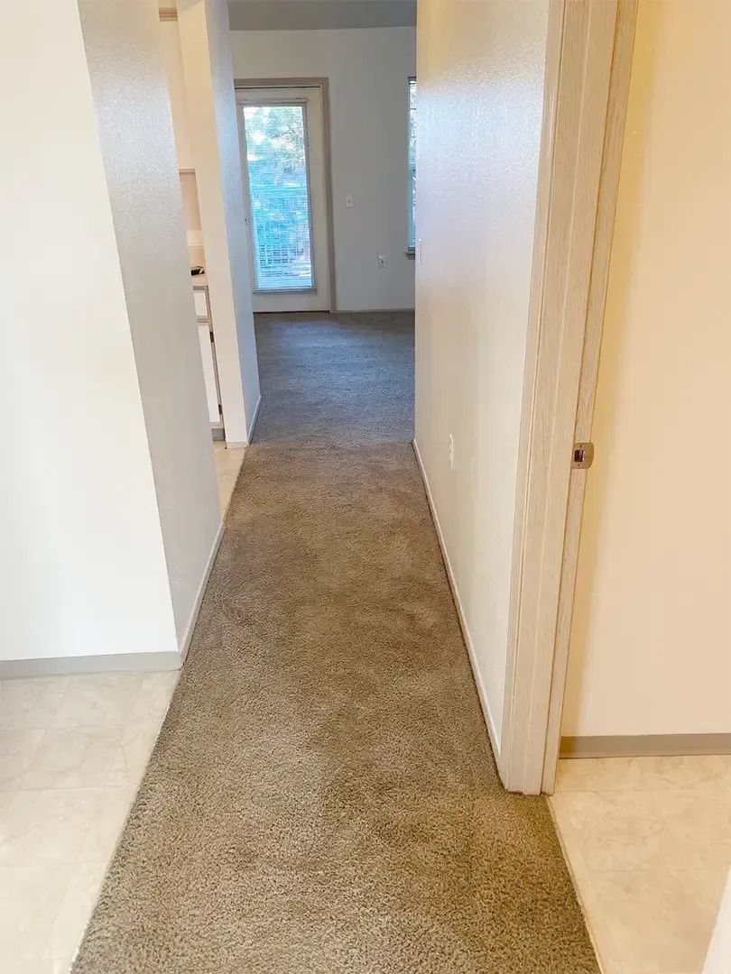 Narrow carpeted hallway with white walls leading to a window at the far end.