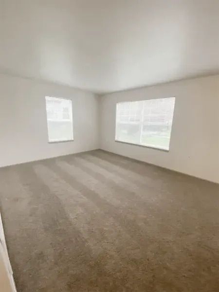 Empty beige carpeted room with two windows and white walls, shown from a corner