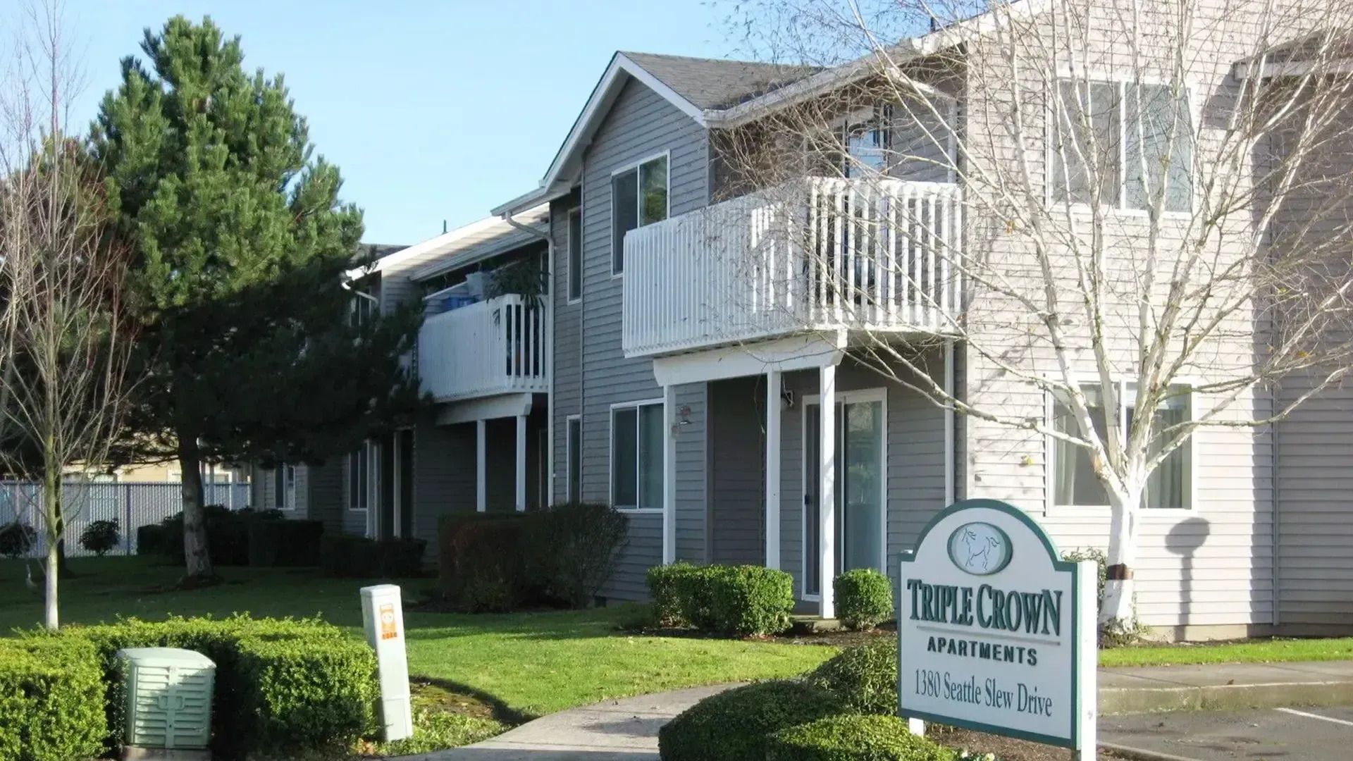Apartment complex exterior with white balconies, trees, and a community sign in the foreground