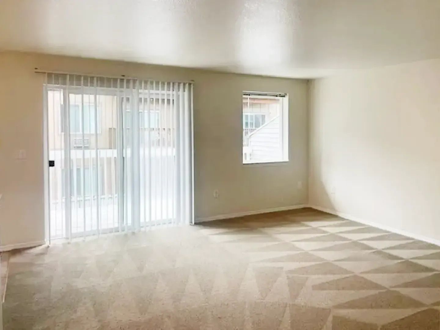 Empty beige living room with carpet, sliding glass doors, and a small window letting in daylight
