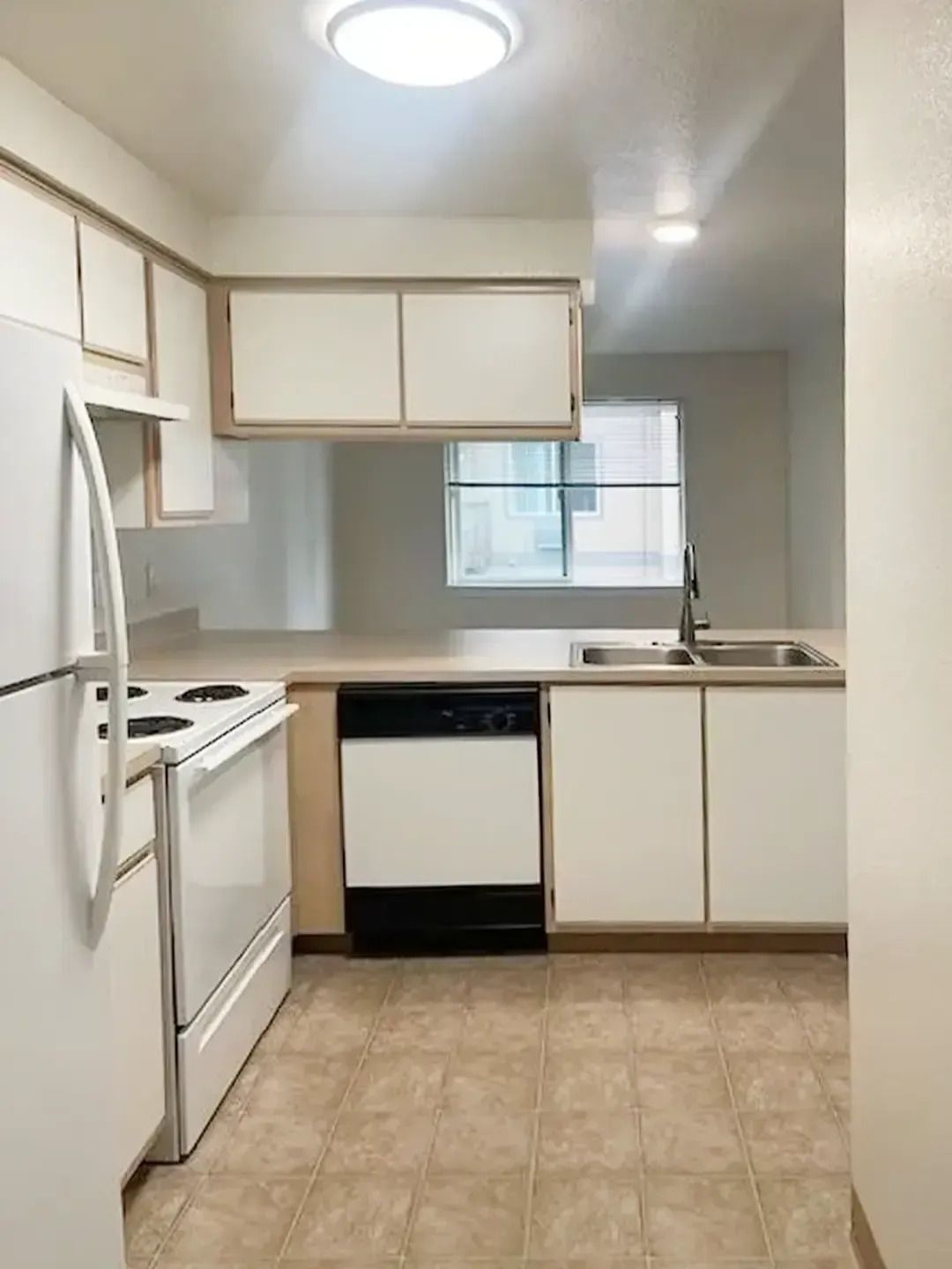 Small kitchen with white appliances, beige cabinets, tiled floor, and a window above the sink.