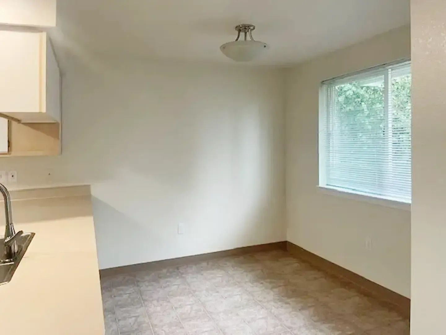 Empty kitchen with white walls, tan carpet, and a window with blinds.