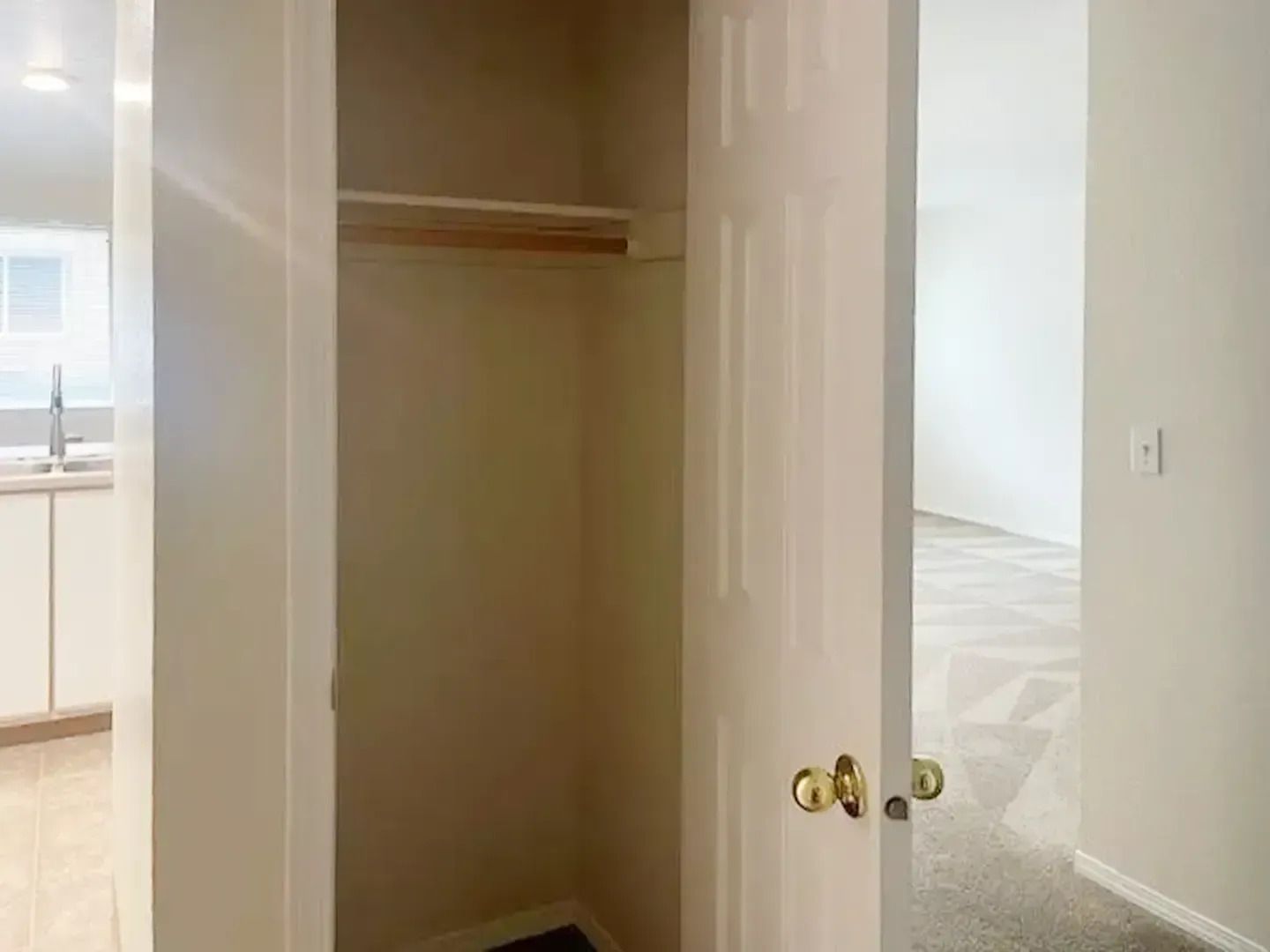 Open closet with hanging rod in a bright empty room, with a doorway and kitchen visible nearby