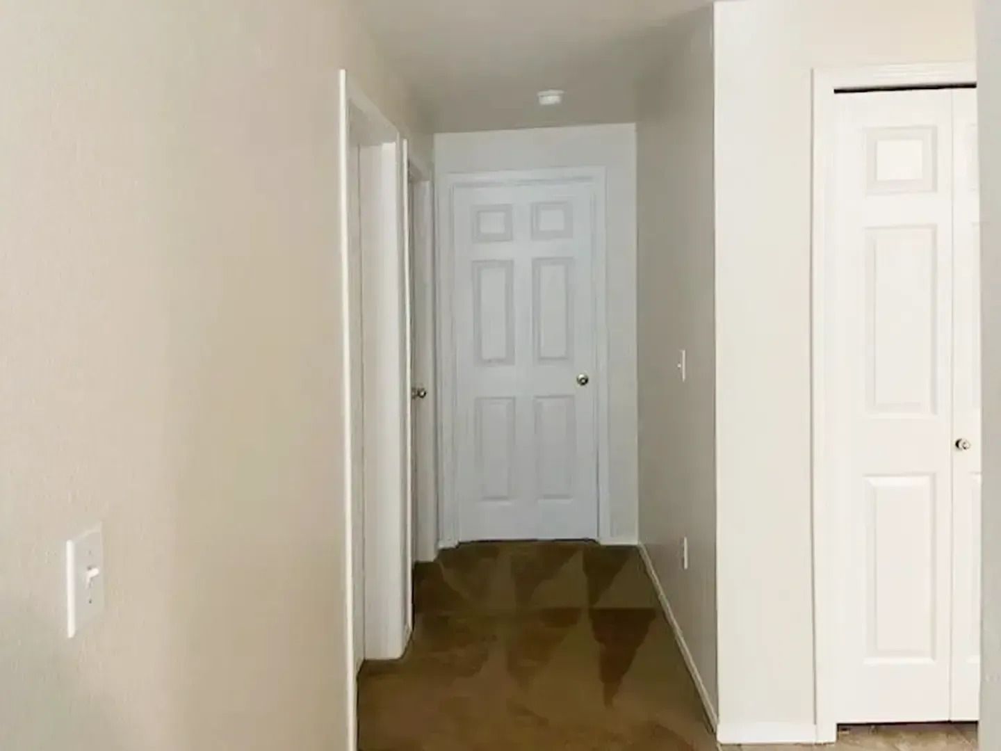 Empty hallway with beige walls, white doors, and brown flooring leading to a closed door.