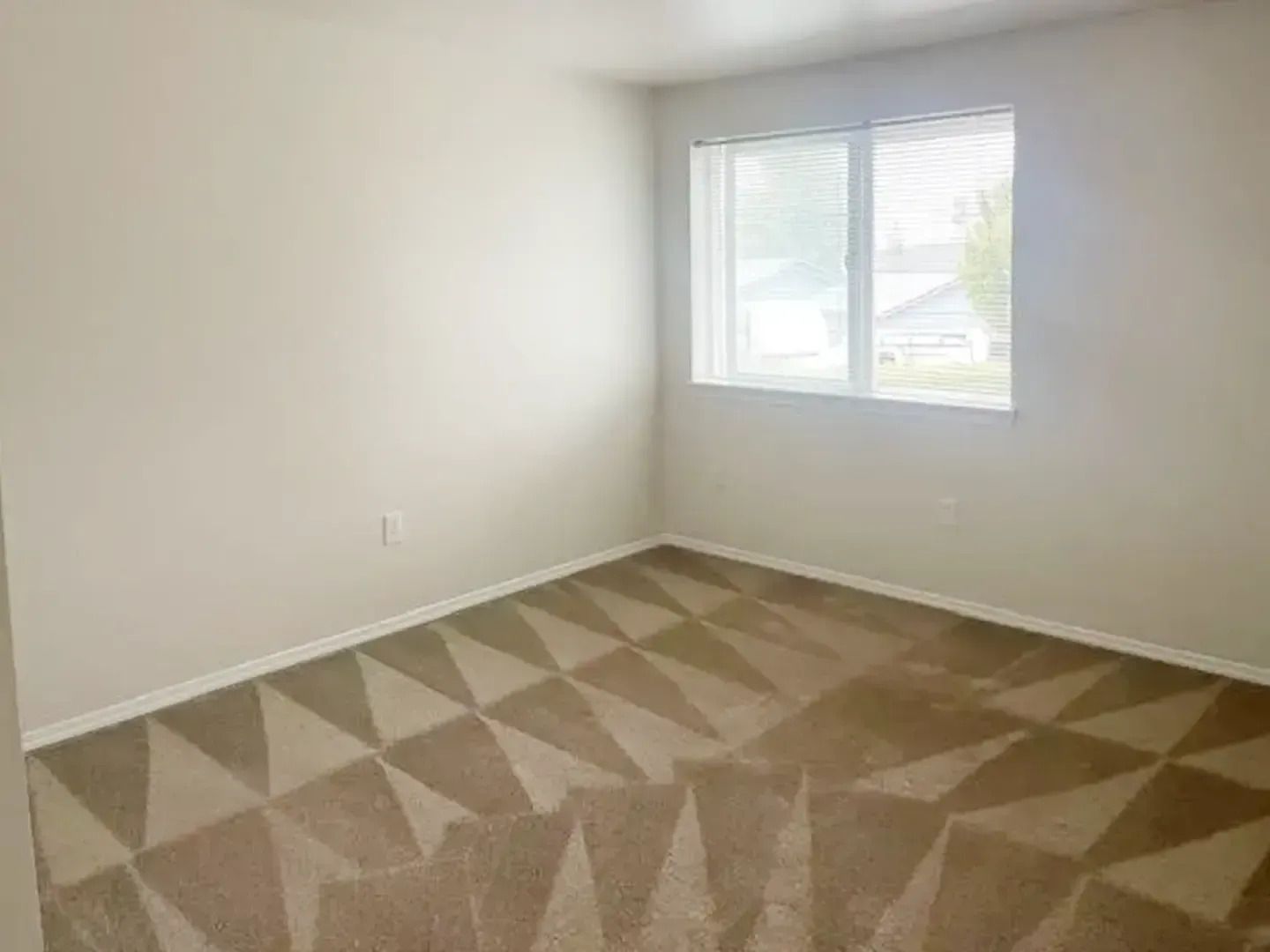 Empty beige bedroom with window and patterned carpet; white walls and natural light.