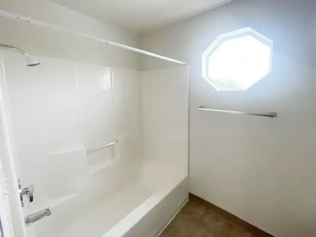 Empty white bathroom with bathtub, wall-mounted towel bar, and octagonal window letting in bright light