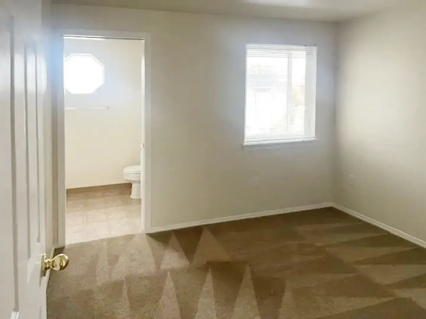 Empty beige bedroom with carpet, window, and open doorway to a bathroom