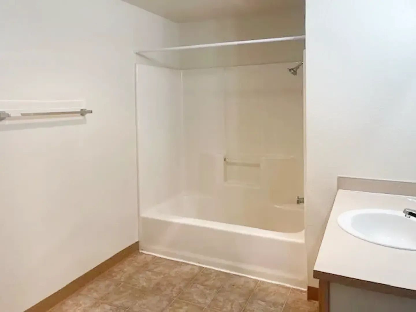 Empty bathroom with shower-tub, vanity sink, towel bar, and beige tile floor.
