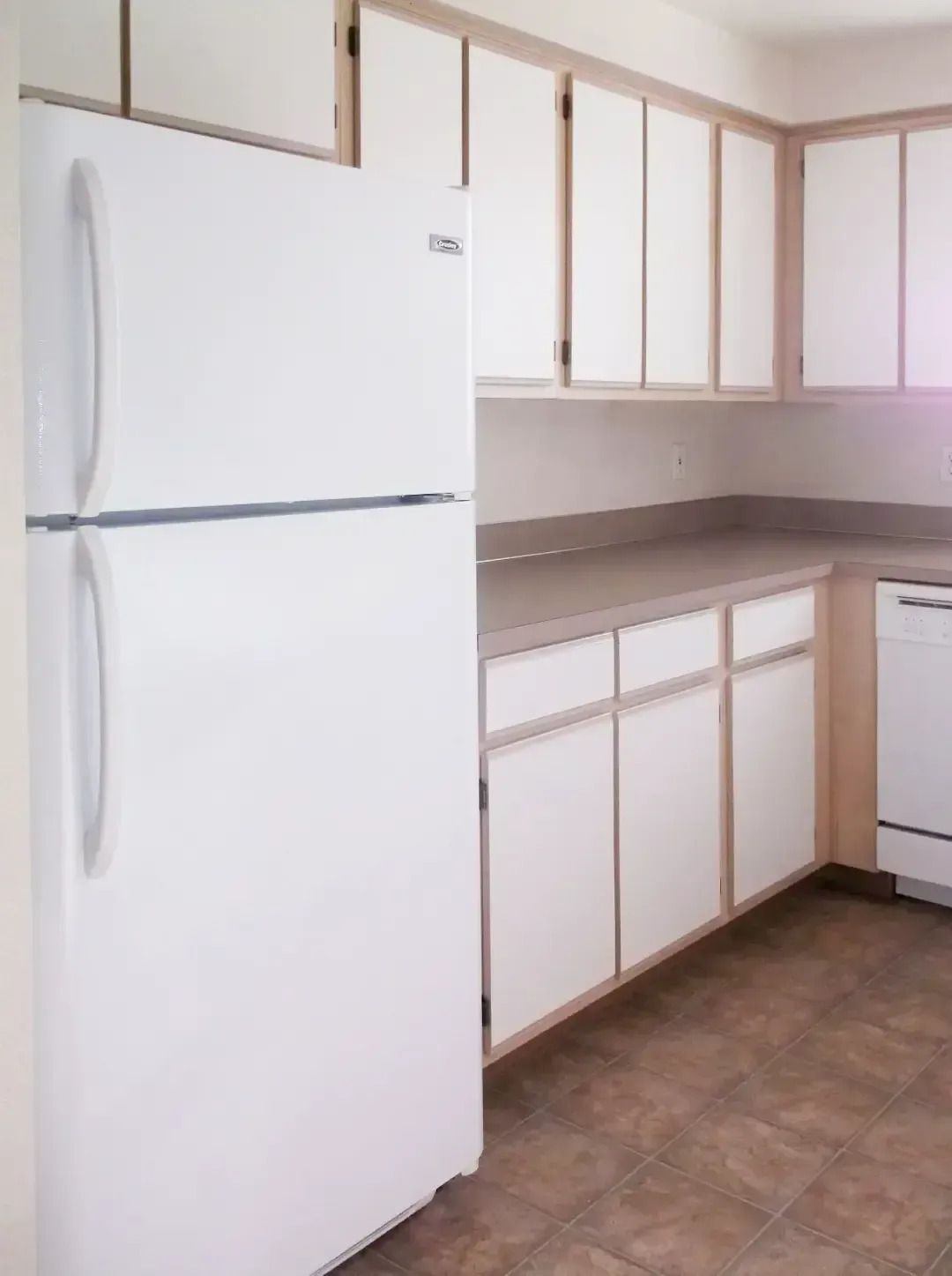 Empty kitchen with white cabinets, white refrigerator, gray countertops, and brown tile floor.