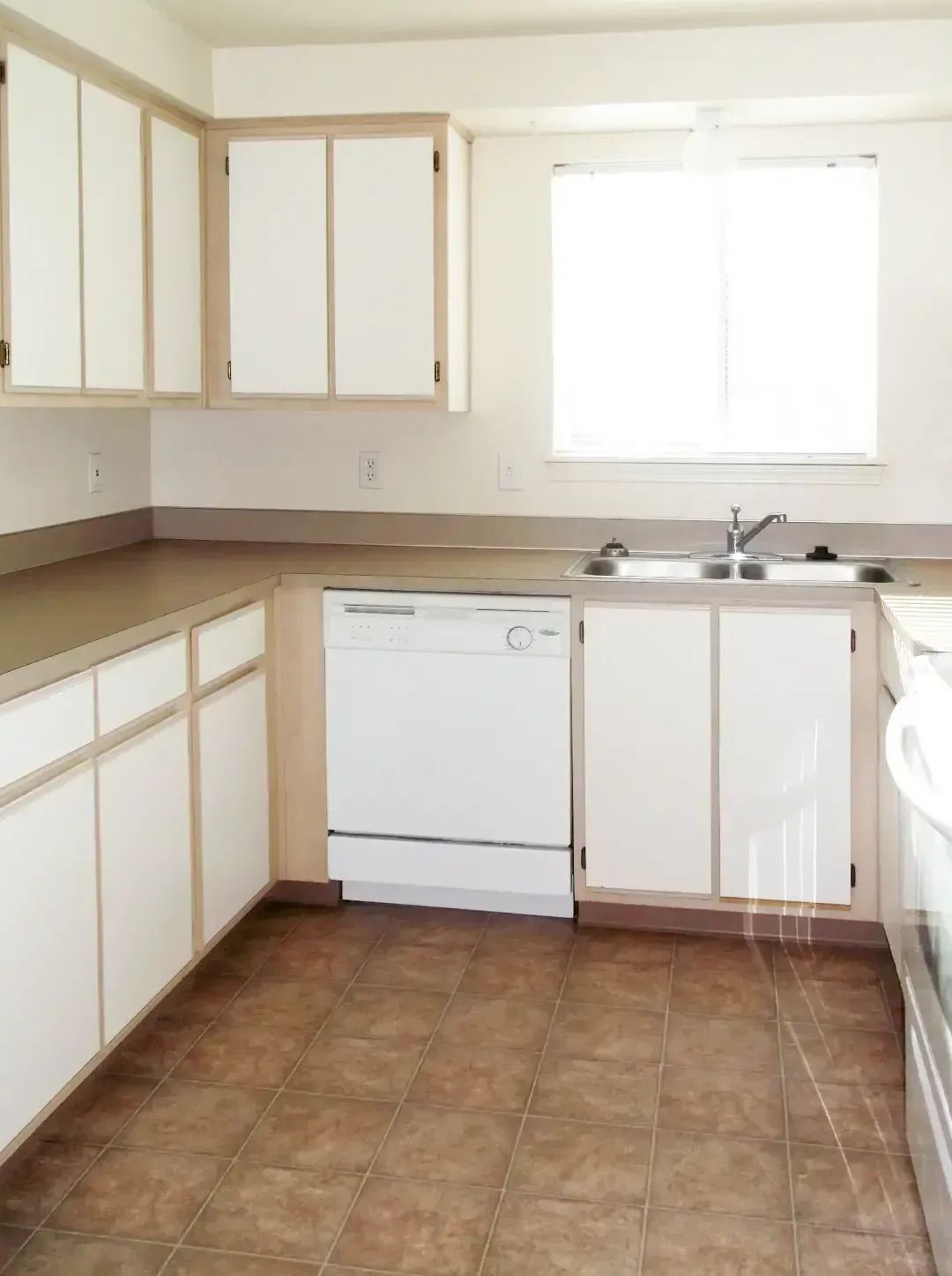 Bright kitchen with white cabinets, tan countertop, dishwasher, sink, and a sunlit window.