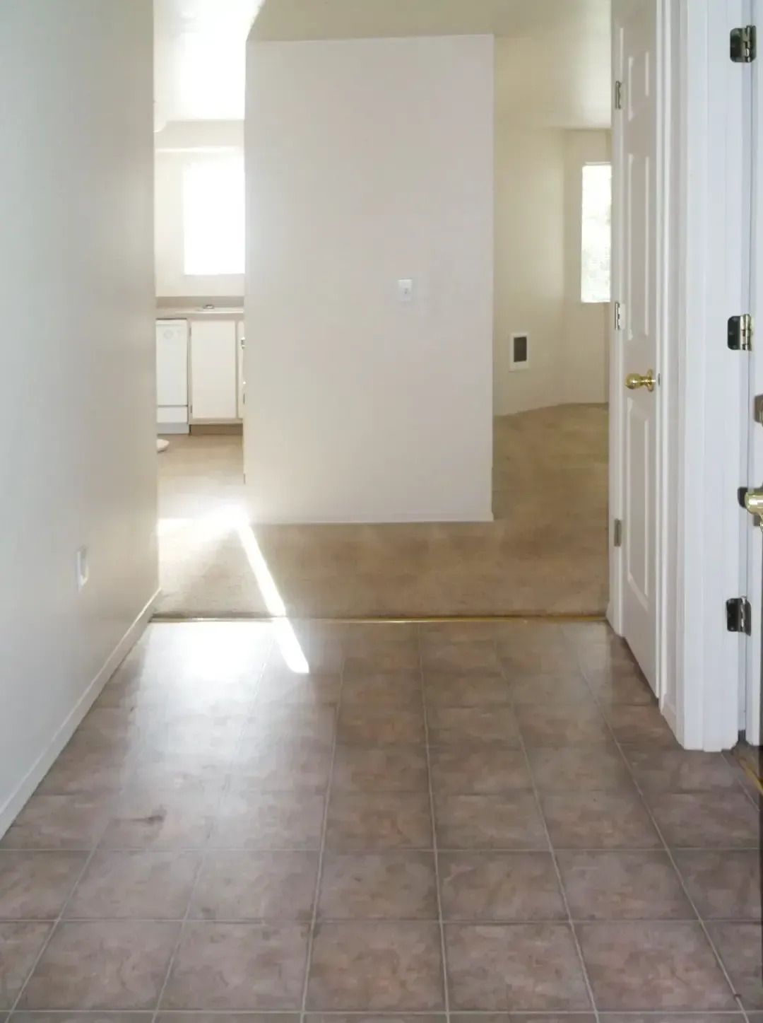 Empty tiled hallway with sunlight streaming through windows into a bright room.