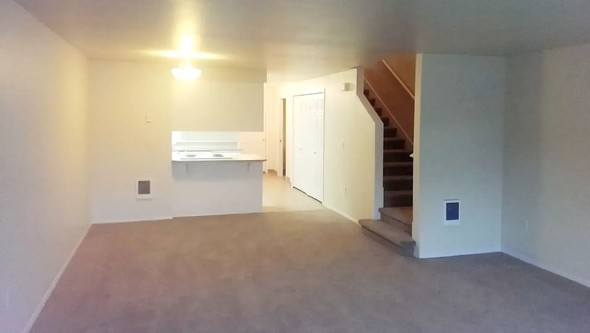 Empty carpeted living room with kitchen nook and staircase leading up on the right.