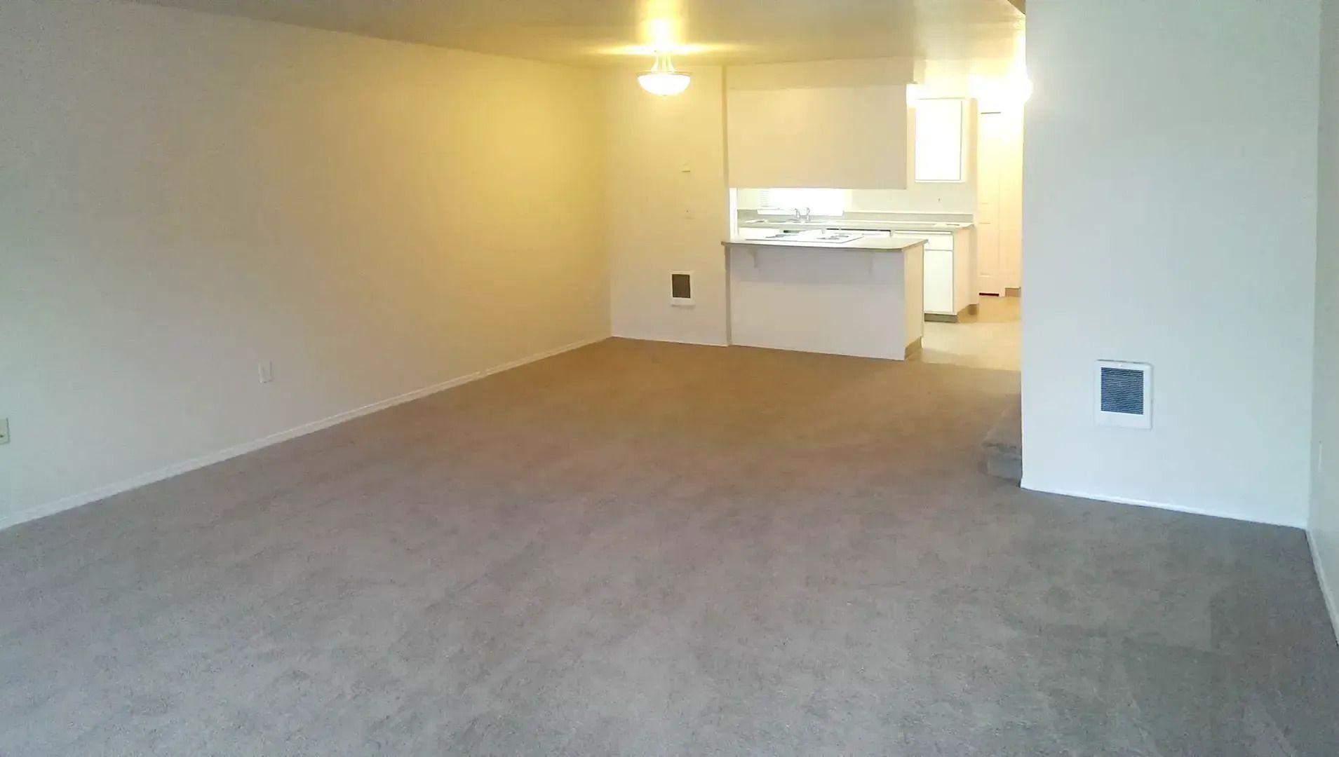 Empty carpeted living room with kitchen in the background and a small wall vent on the right