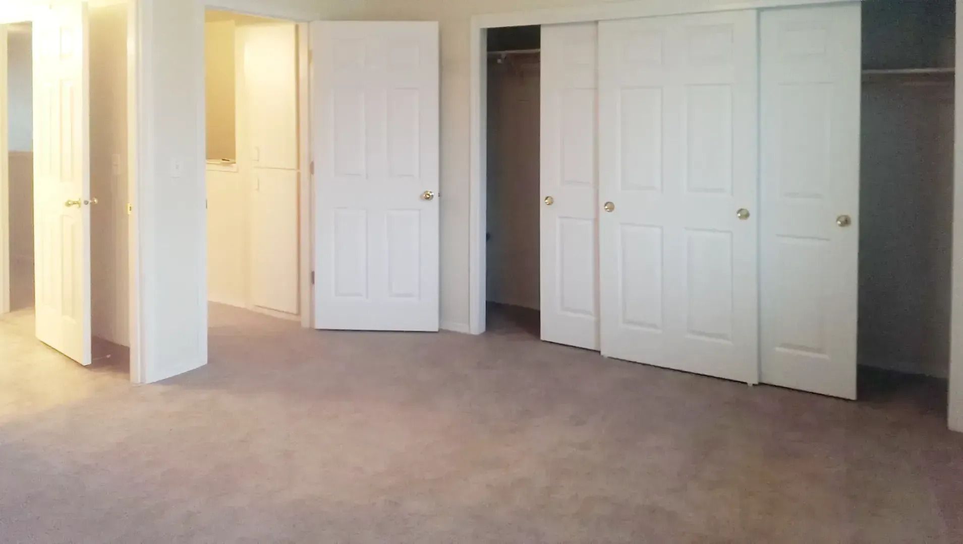 Empty beige-carpeted bedroom with white double closet doors and an open doorway to a lit hall
