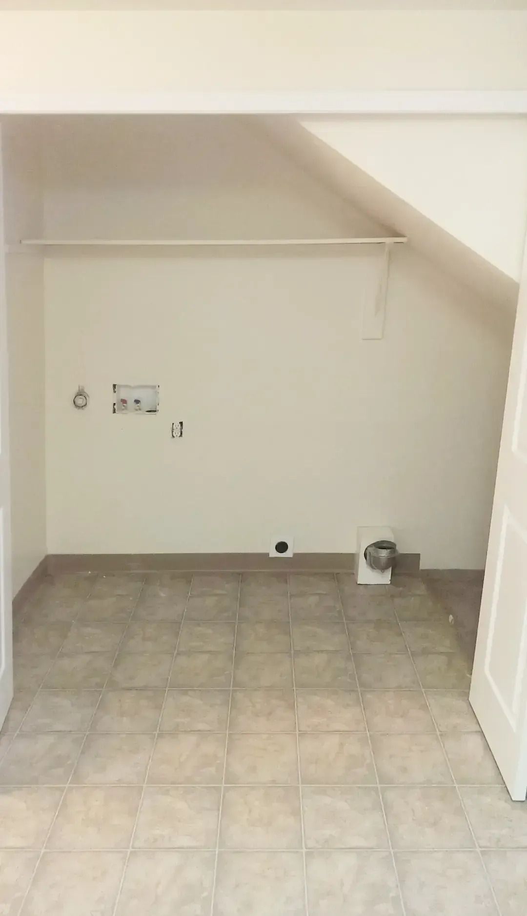 Empty small room with tiled floor, slanted ceiling, and a wall shelf near utility hookups.