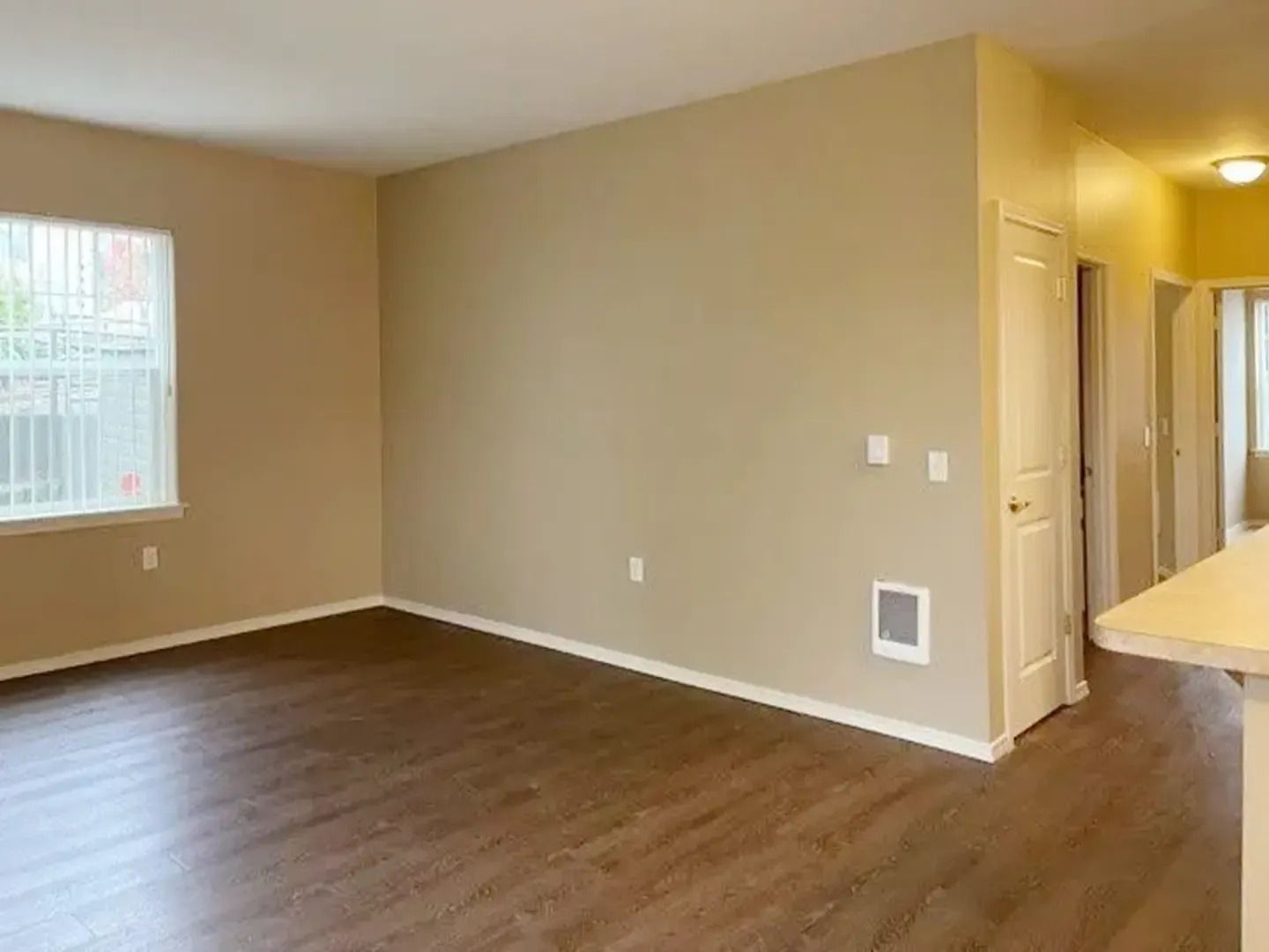 Empty beige living room with wood floors, a small window, and a kitchen counter on the right