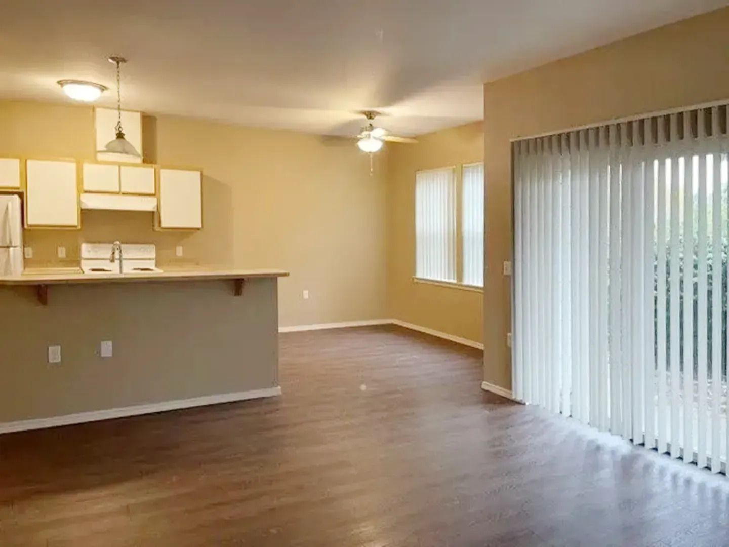Empty living room with kitchen bar, wood floors, beige walls, and vertical blinds.