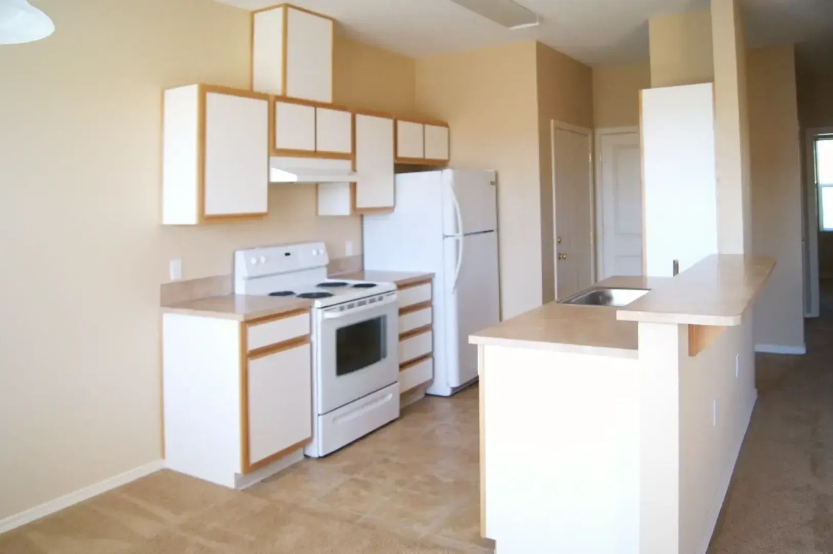 Empty beige kitchen with white appliances and a small breakfast bar.