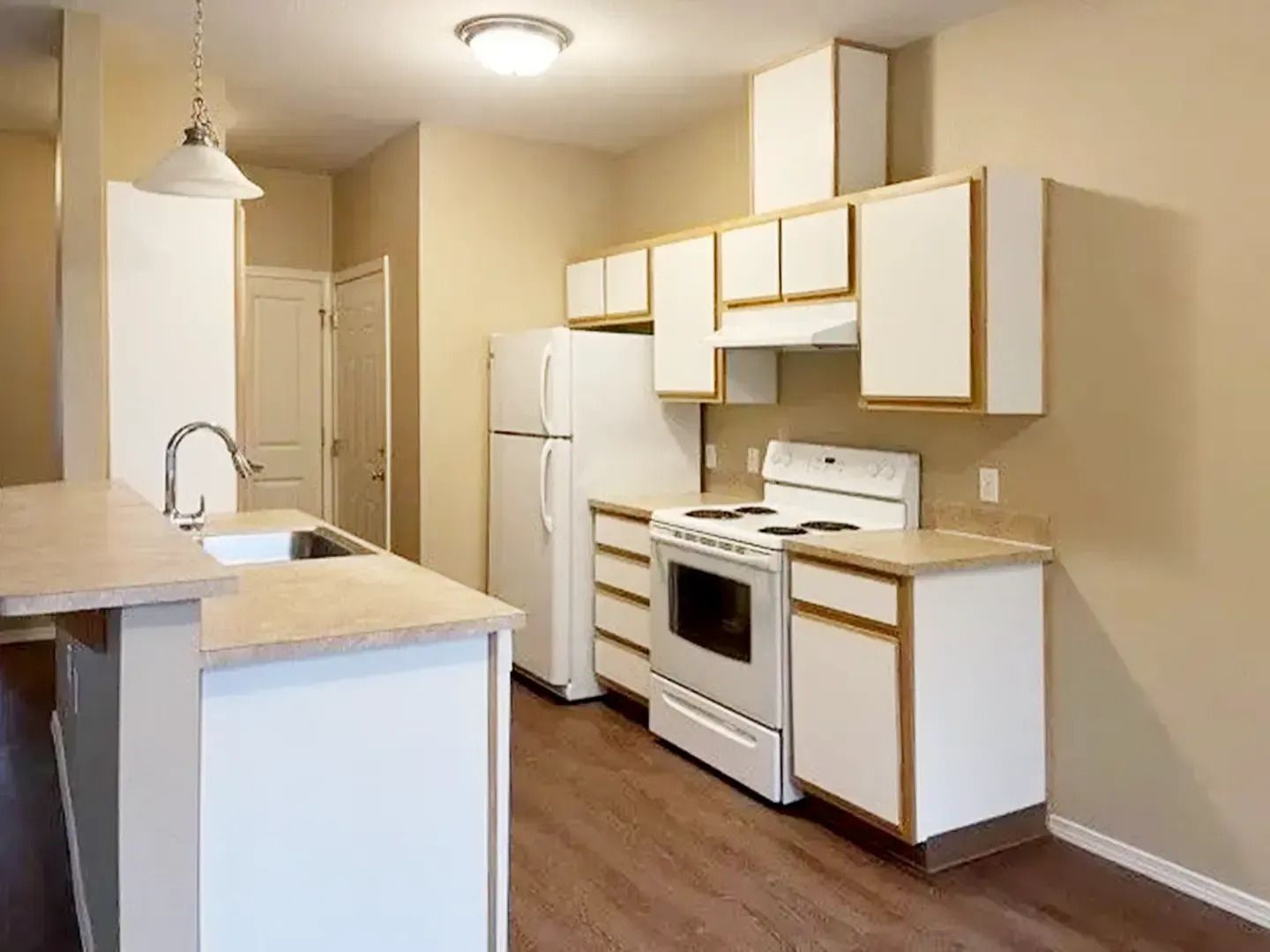 Small beige kitchen with white cabinets, stove, refrigerator, and breakfast bar lighting.