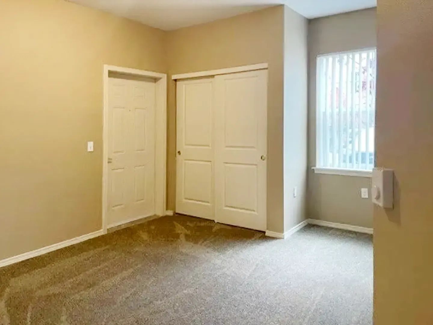 Empty beige bedroom with carpet, two white closet doors, and a window with vertical blinds