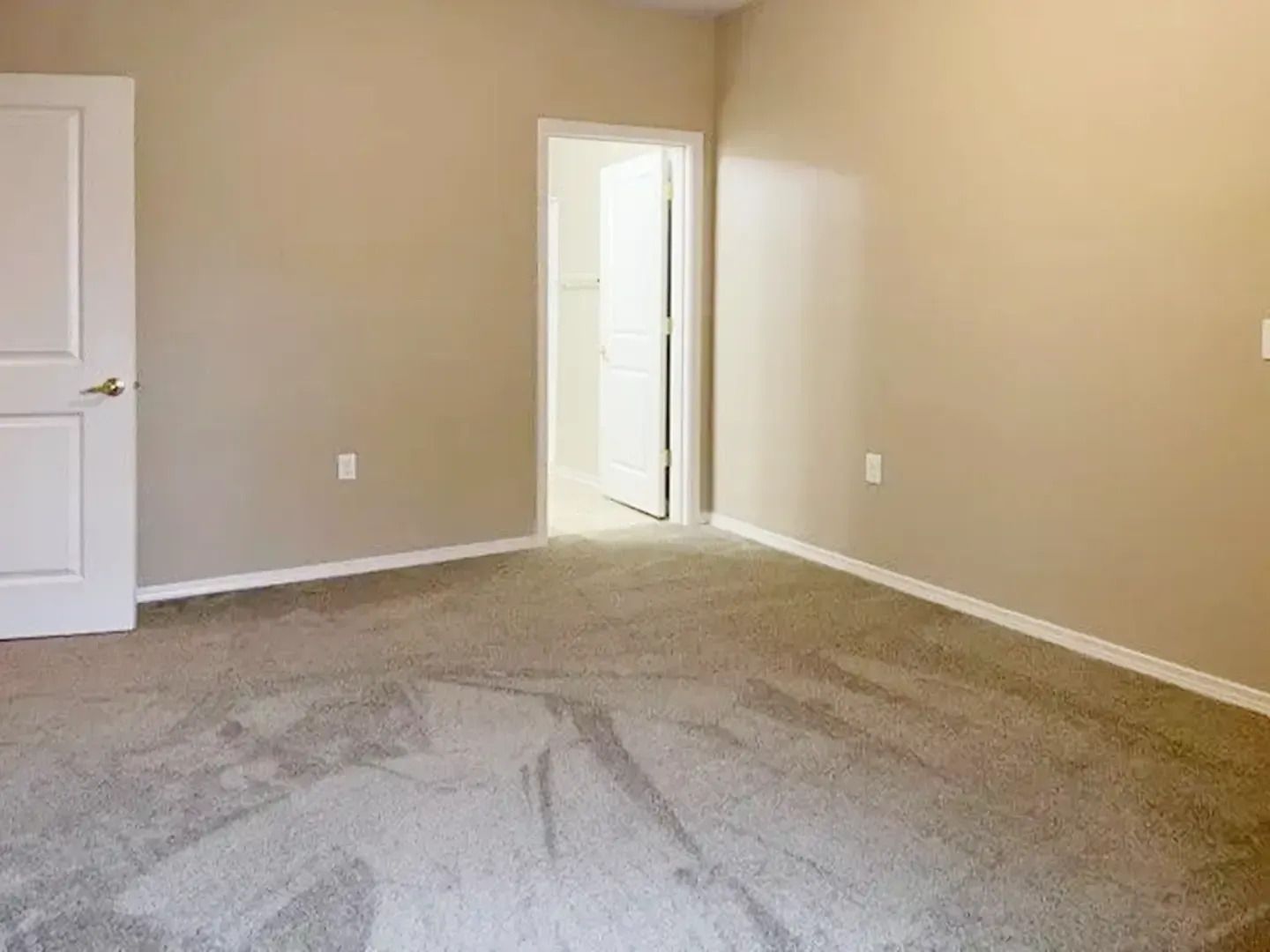 Empty beige bedroom with carpet, white door, and open doorway to a lit hall