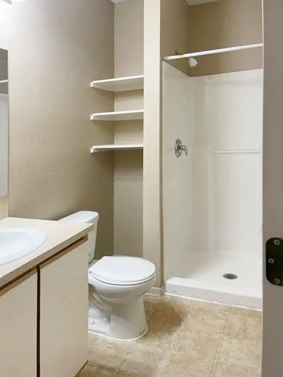 Small beige bathroom with toilet, shower stall, vanity, and built-in shelves