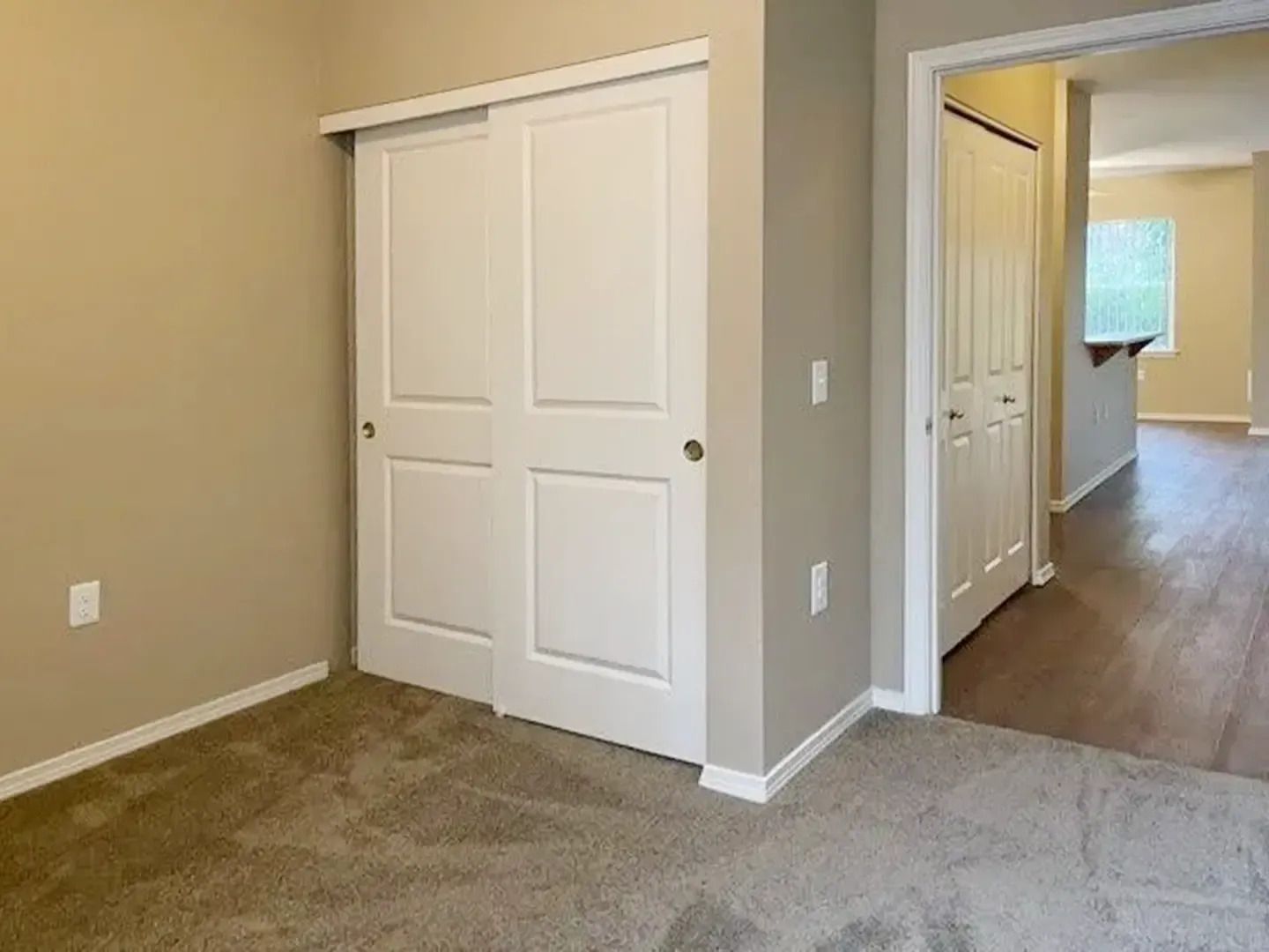 Empty beige bedroom with white closet doors and an open doorway to a hallway with wood flooring