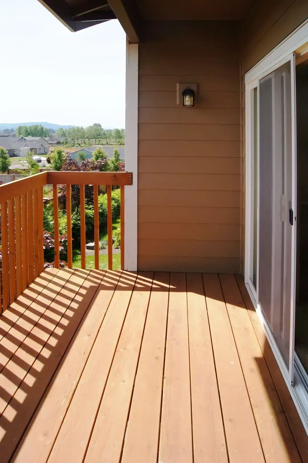 Sunlit wooden deck with railing, sliding glass door, and view of houses beyond