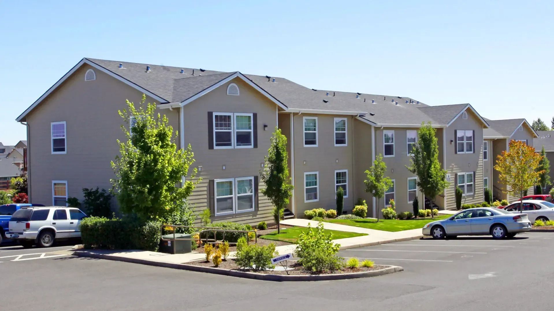 Apartment complex exterior with parked cars, trees, and landscaped entrance in daylight