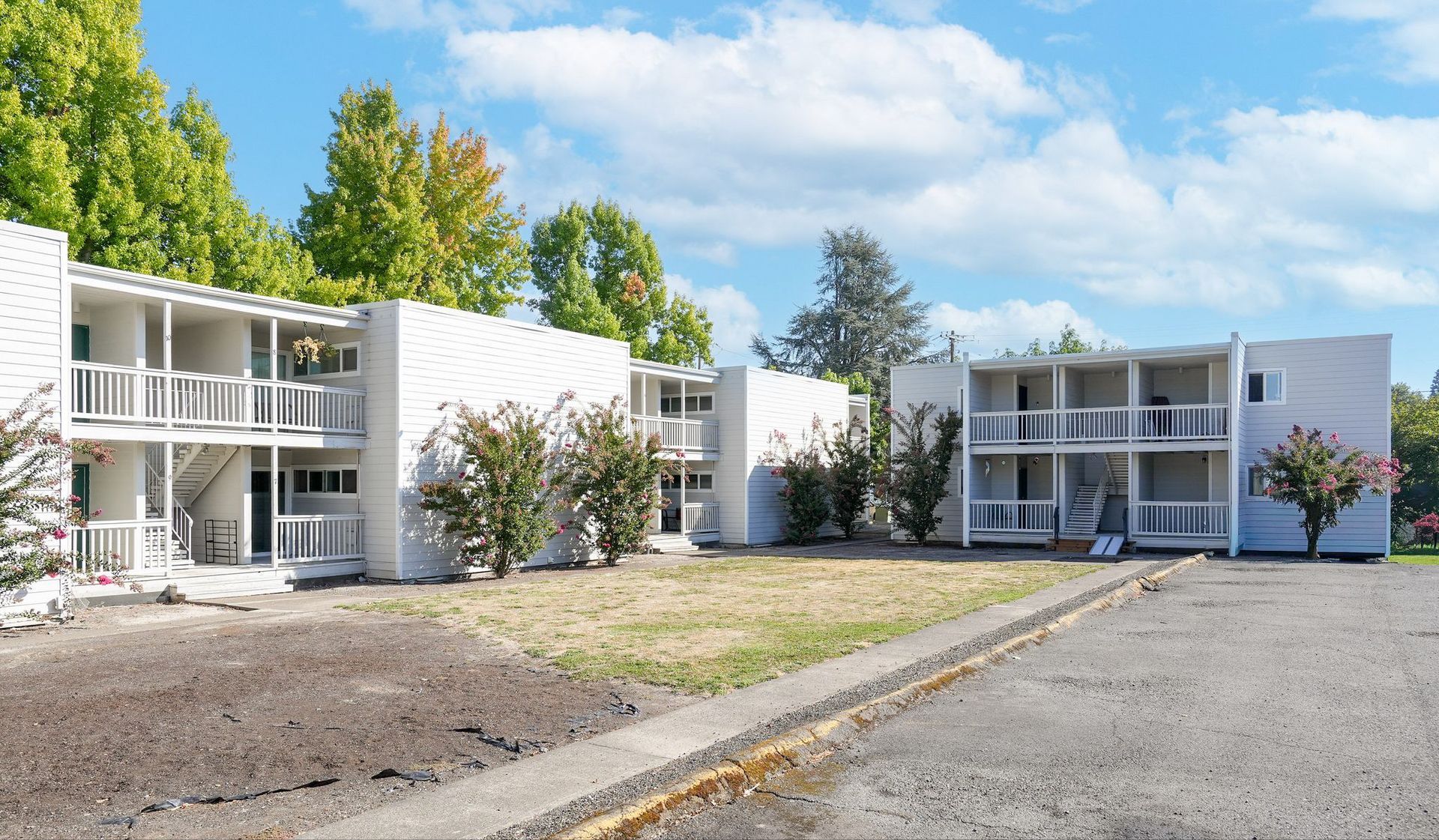 Two white apartment buildings with balconies beside a gravel lot and sparse trees under a blue sky