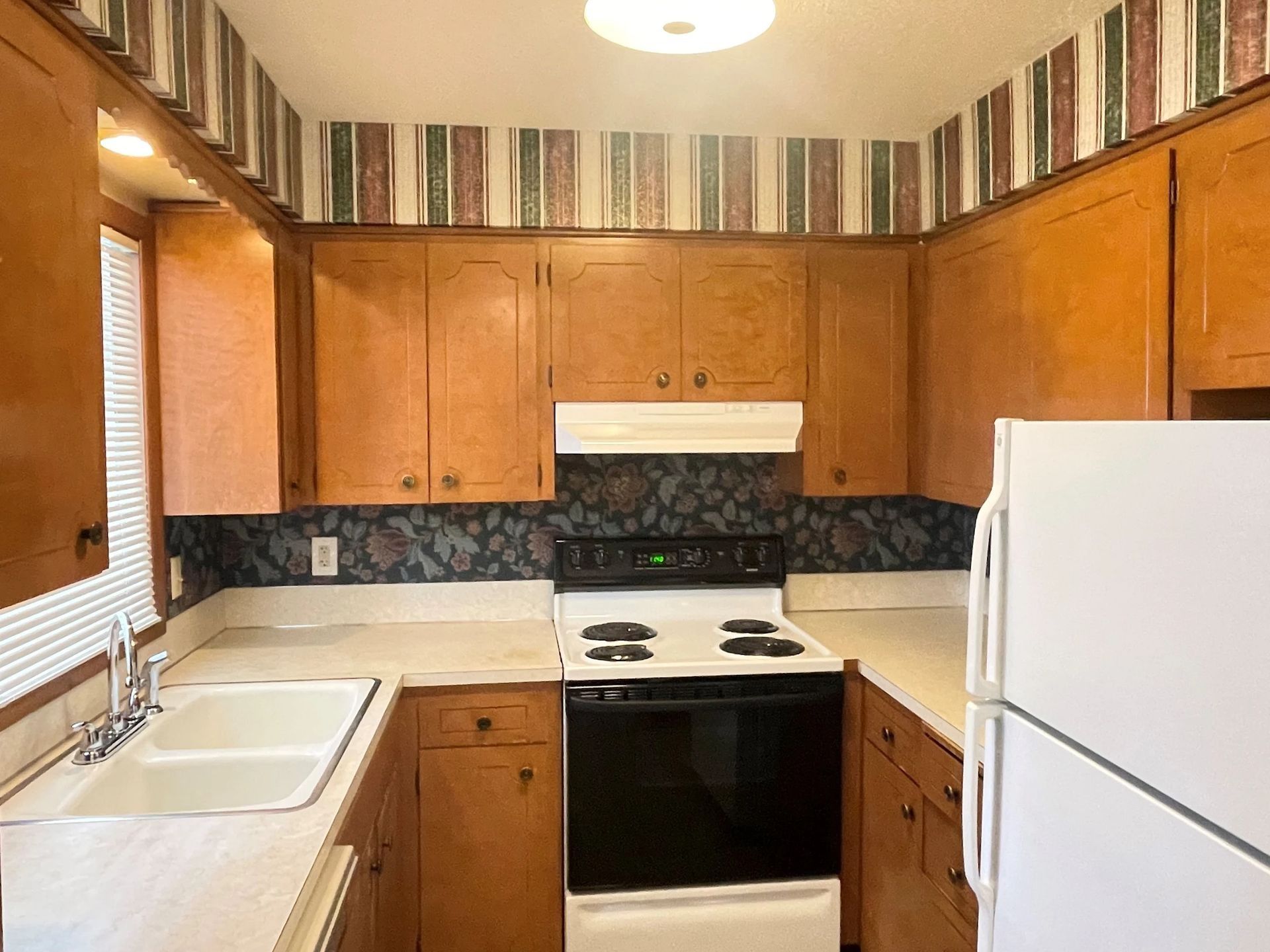 Small kitchen with wood cabinets, white appliances, striped wallpaper, and a double sink.