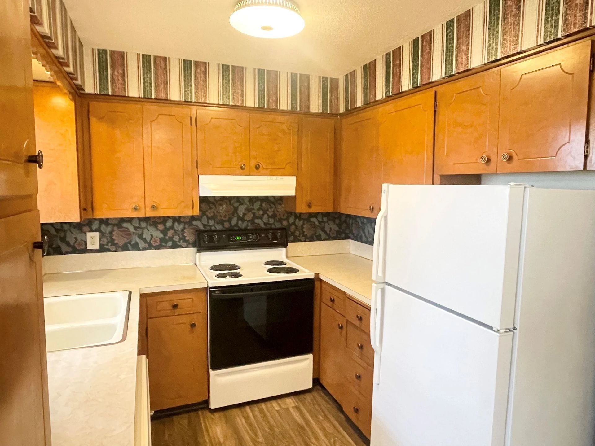 Small kitchen with wood cabinets, white appliances, striped wallpaper, and a black stove.