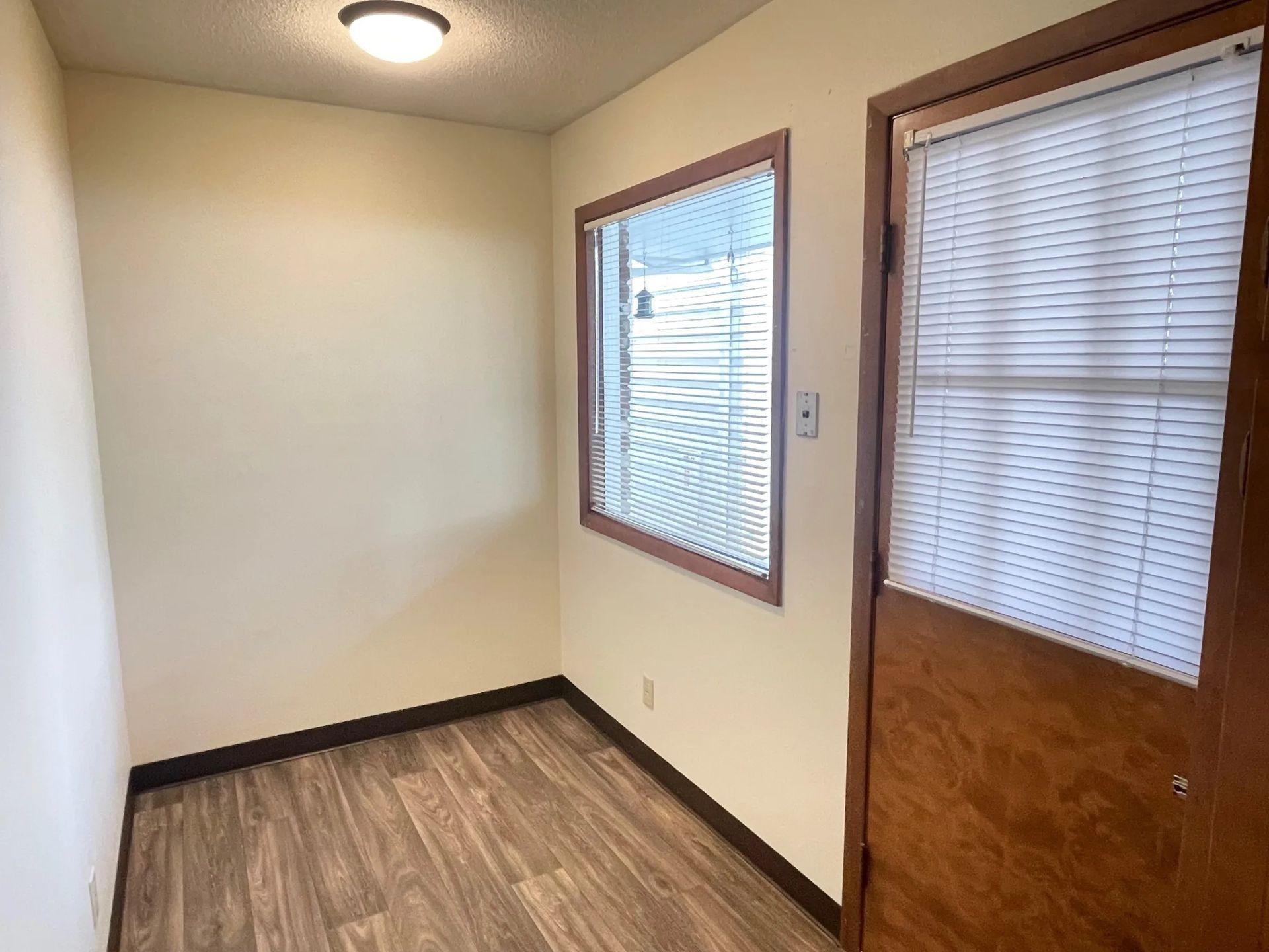 Empty room with wood-look floor, beige walls, and two windows beside a wooden door with blinds.