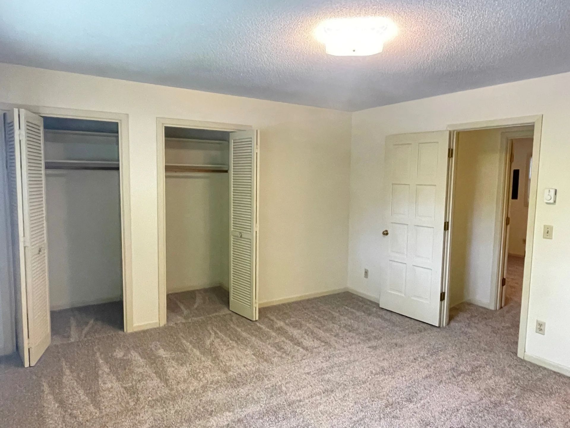 Empty beige bedroom with carpet, three closets, and an open door to a hallway.
