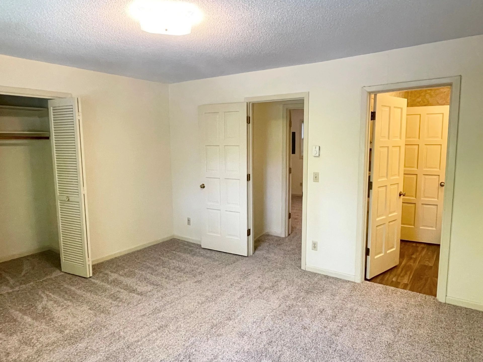 Empty beige carpeted bedroom with open closet and two open doors leading to hall and bathroom.