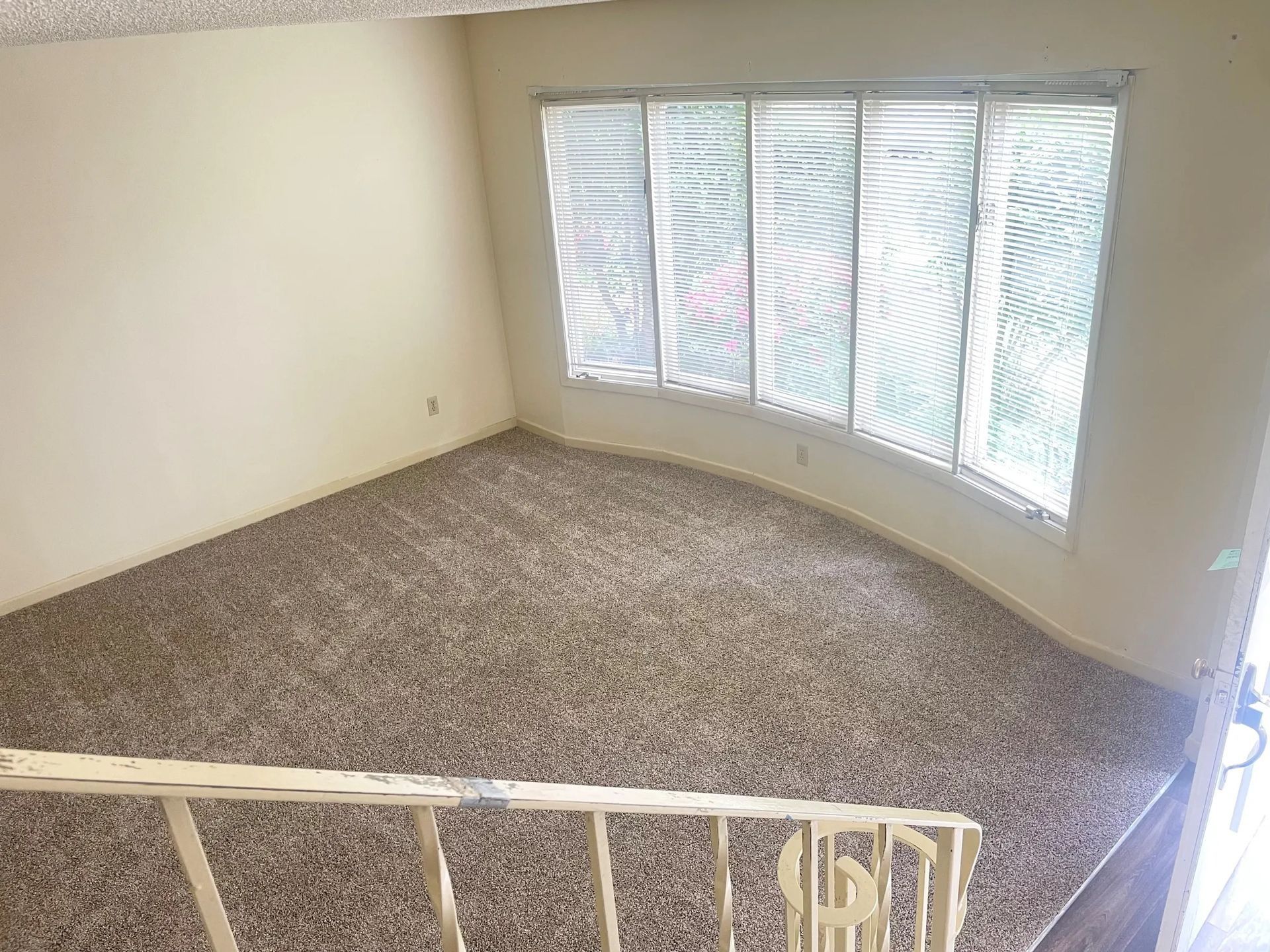 Empty carpeted room with beige walls and large windows with white blinds