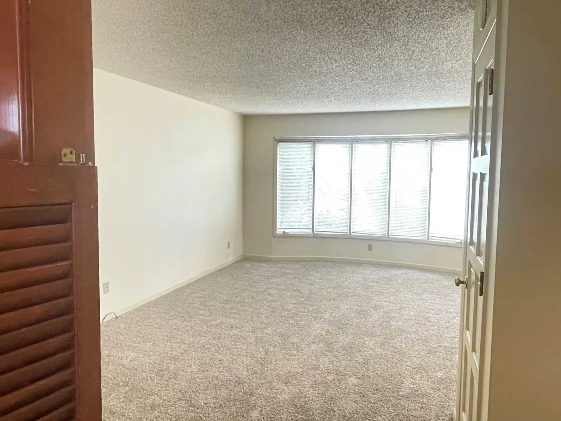 Empty carpeted living room with white walls and a large window with shutters