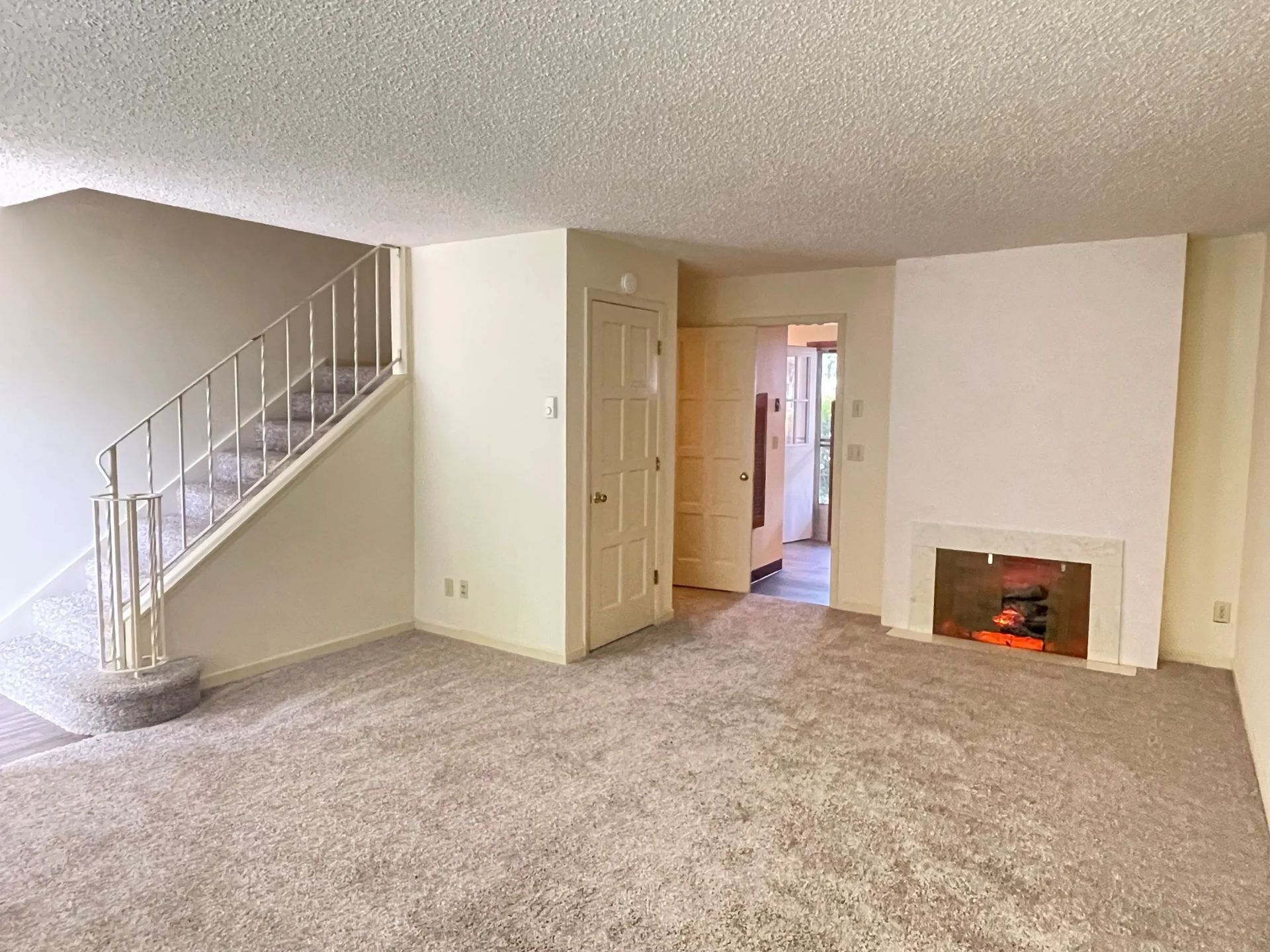 Empty living room with carpet, staircase, doorway, and a small fireplace on the right.